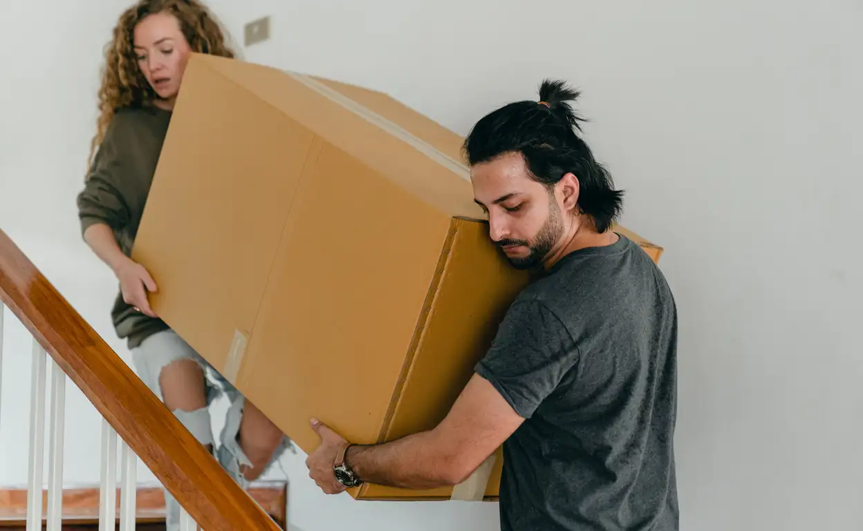 Twee studenten zijn aan het verhuizen en tillen een grote verhuisdoos een trap op.