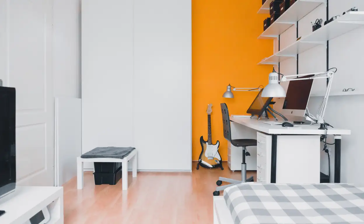 Een studentenkamer met een oranje muur en een gitaar tegen de muur
