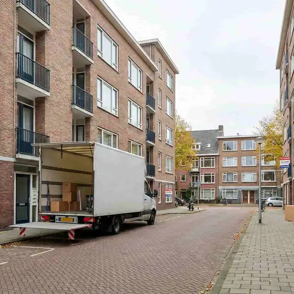 Verhuiswagen voor appartementencomplex, jong stel draagt dozen, 'Te Koop Verkocht' bord in raam.