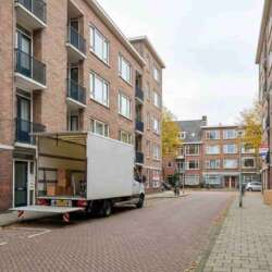 Verhuiswagen voor appartementencomplex, jong stel draagt dozen, 'Te Koop Verkocht' bord in raam.